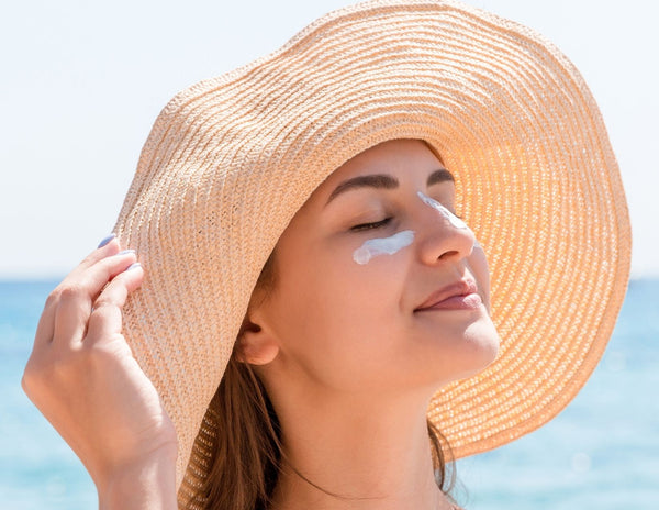 Caucasian woman with sunscreen lotion on her cheek wearing a floppy sun hat with the ocean in the background